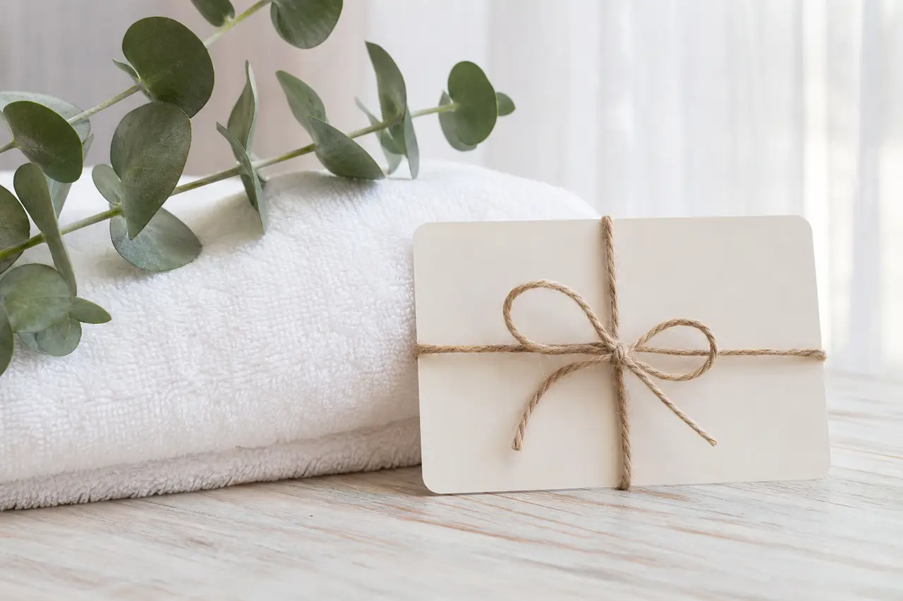 A plain gift card tied with twine beside a folded white towel and a sprig of eucalyptus on a pale wooden table