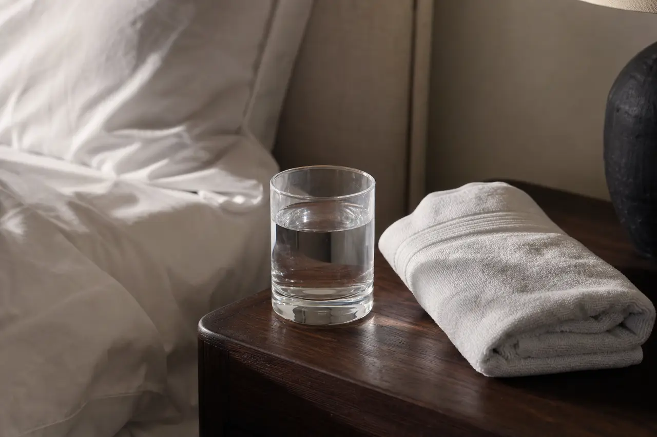 A glass of water and a folded towel on a bedside table in flat morning light