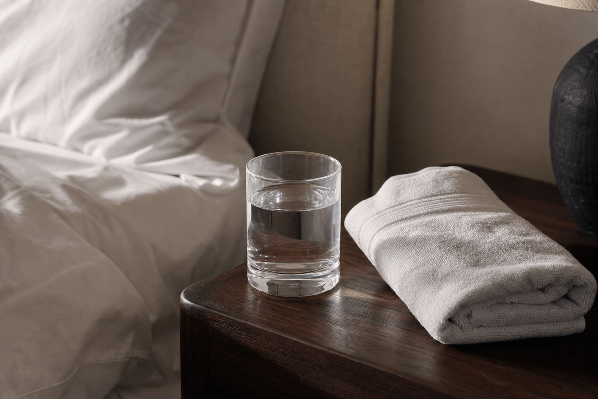 A glass of water and a folded towel on a bedside table in flat morning light
