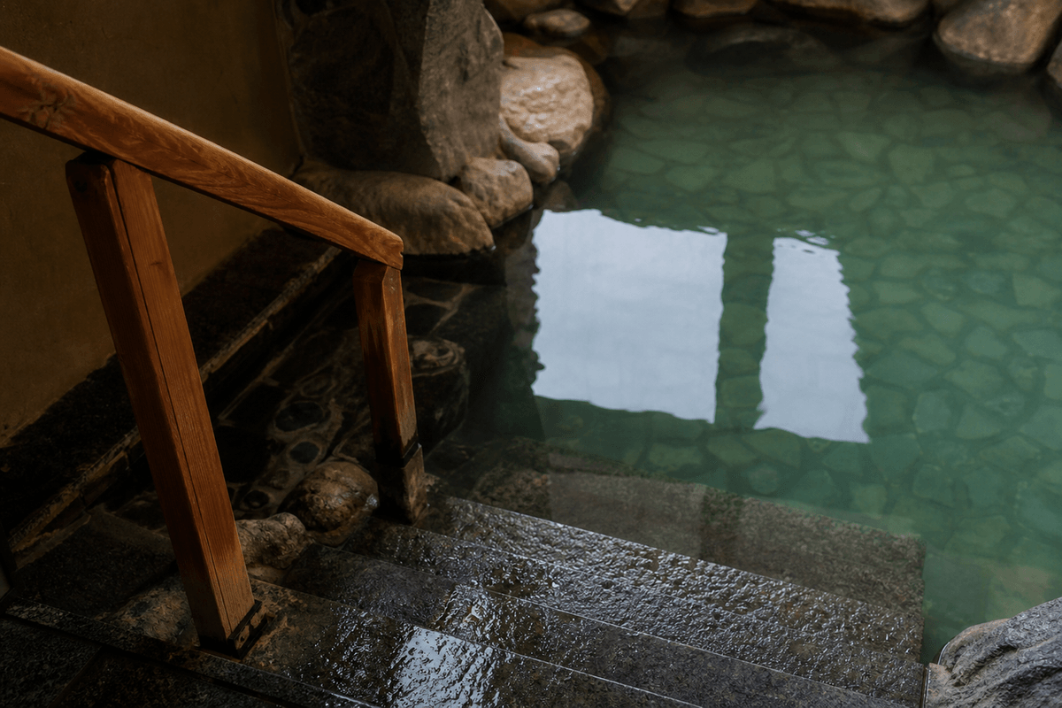 A wooden handrail beside wet stone steps leading down into a calm green plunge pool