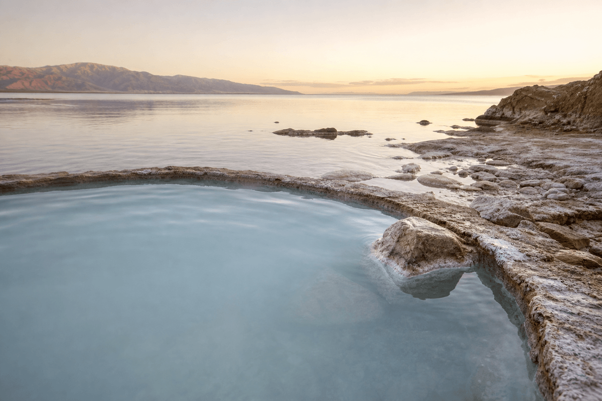 A pale mineral pool at the edge of a salt lake at dawn, crusted white shoreline and hills in the distance
