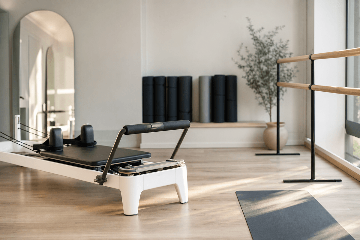 A bright studio with a reformer machine, a ballet barre and rolled mats along the wall