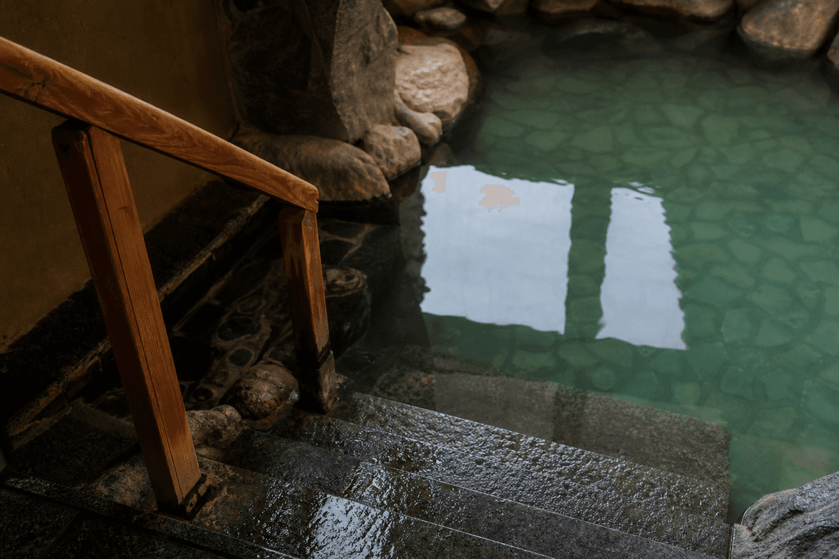 A wooden handrail beside wet stone steps leading down into a calm green plunge pool