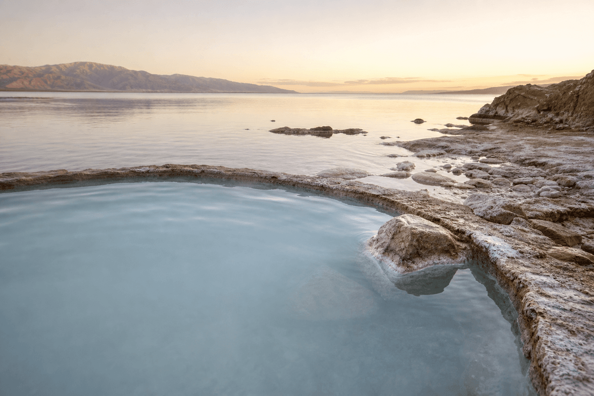 A pale mineral pool at the edge of a salt lake at dawn, crusted white shoreline and hills in the distance