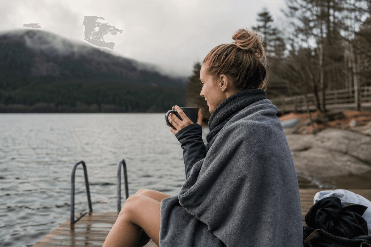 A person wrapped in a towel and warm layers holding a hot drink after a cold plunge outdoors