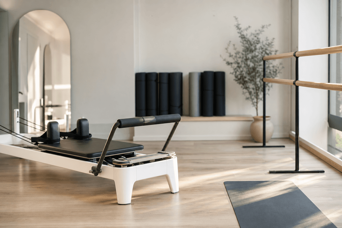 A bright studio with a reformer machine, a ballet barre and rolled mats along the wall